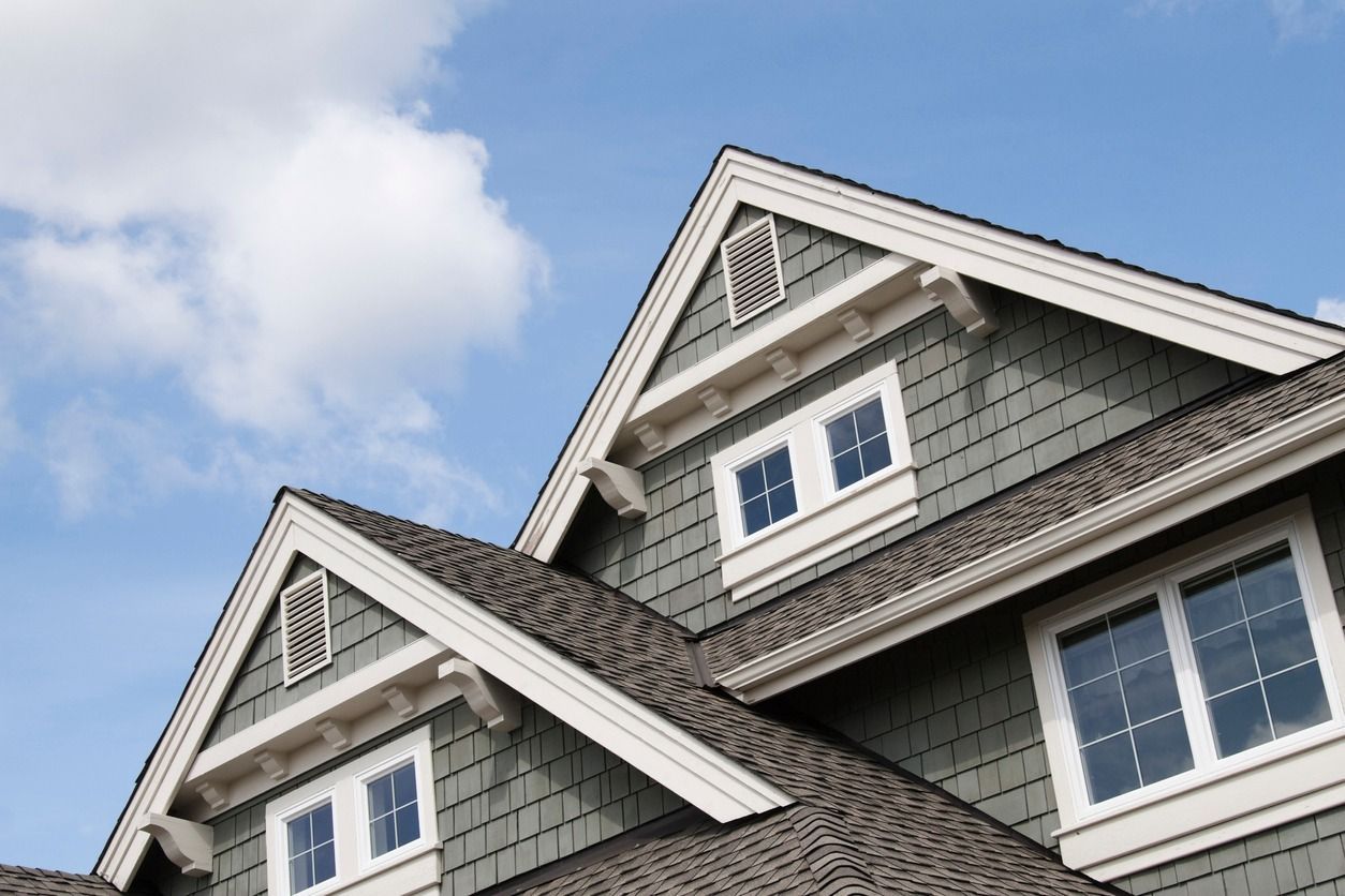 Close-up of a house's upper story with a gray exterior, white trim, windows, and a brown roof against a blue sky.