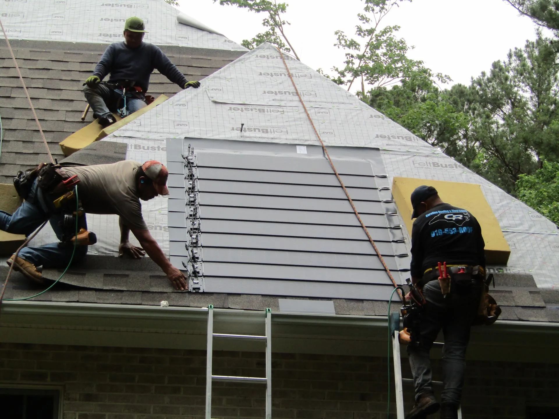 Construction workers installing a large vent on a house roof. They are wearing safety harnesses and are using a ladder.