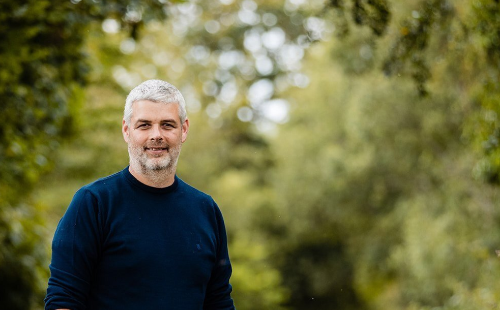 a man with a beard is standing in front of a forest .