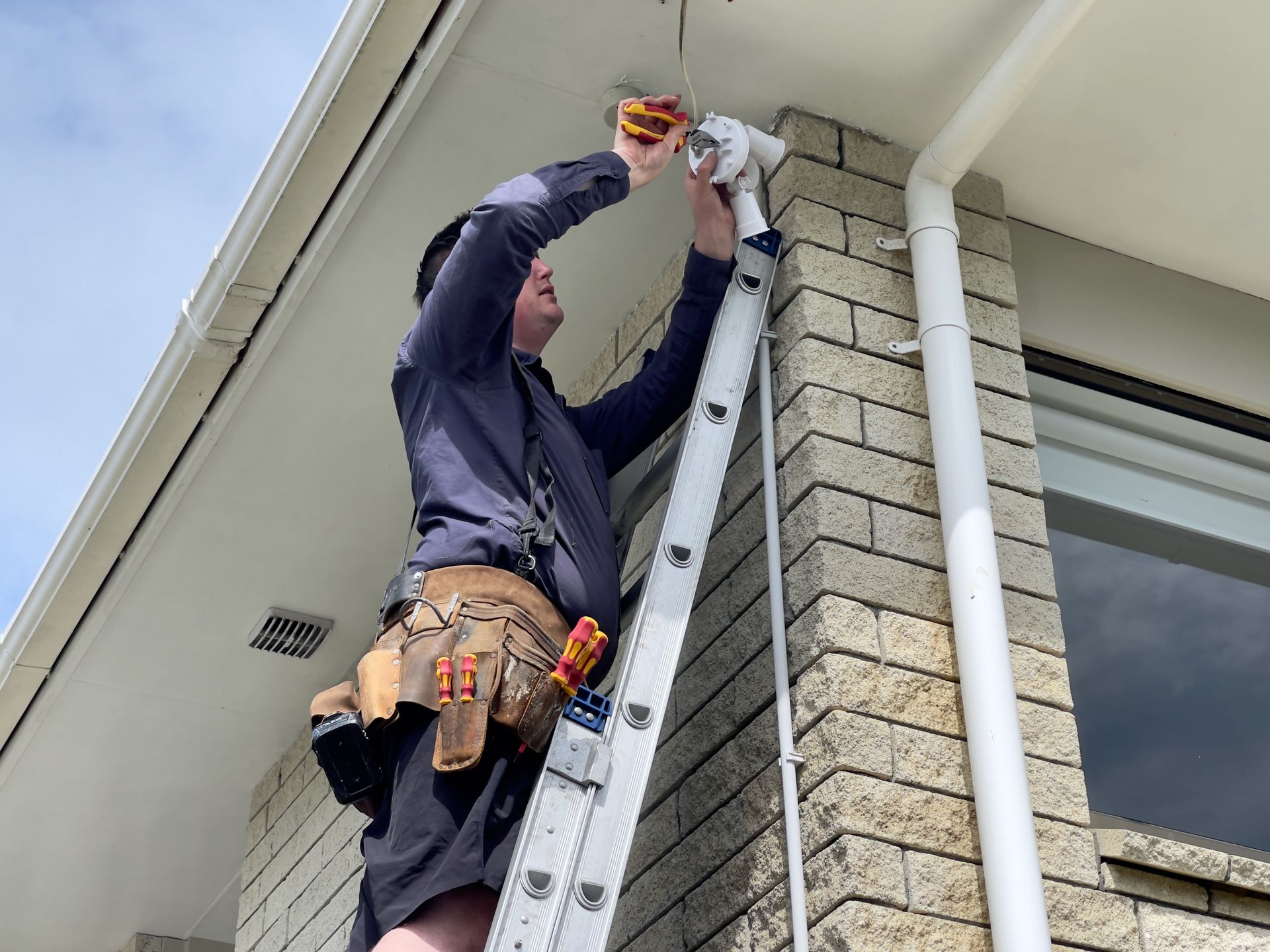 Worker on a ladder repairing the roof edge of a house beside a brick wall.