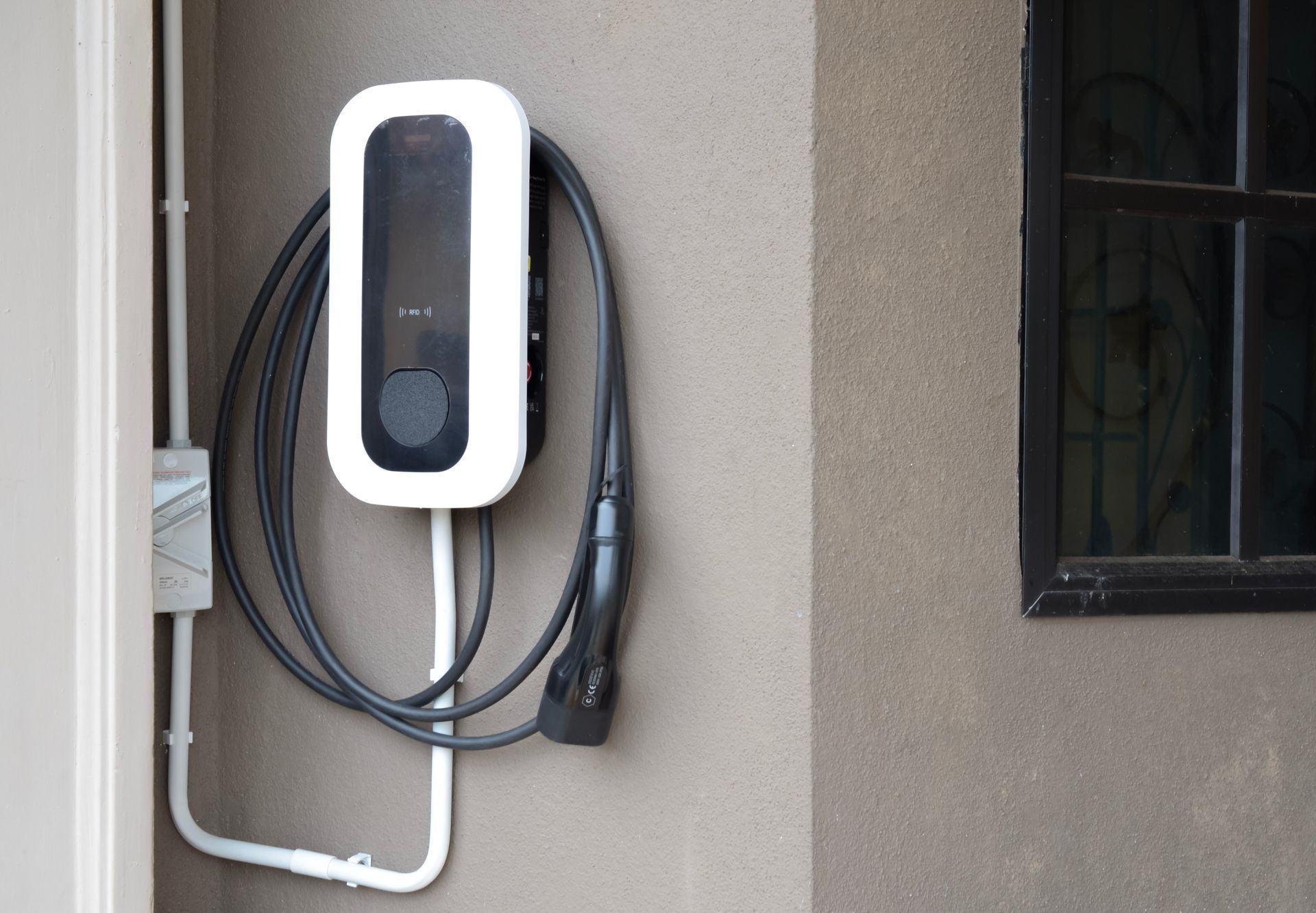 Wall-mounted EV charger with coiled cable beside a house window and doorframe.
