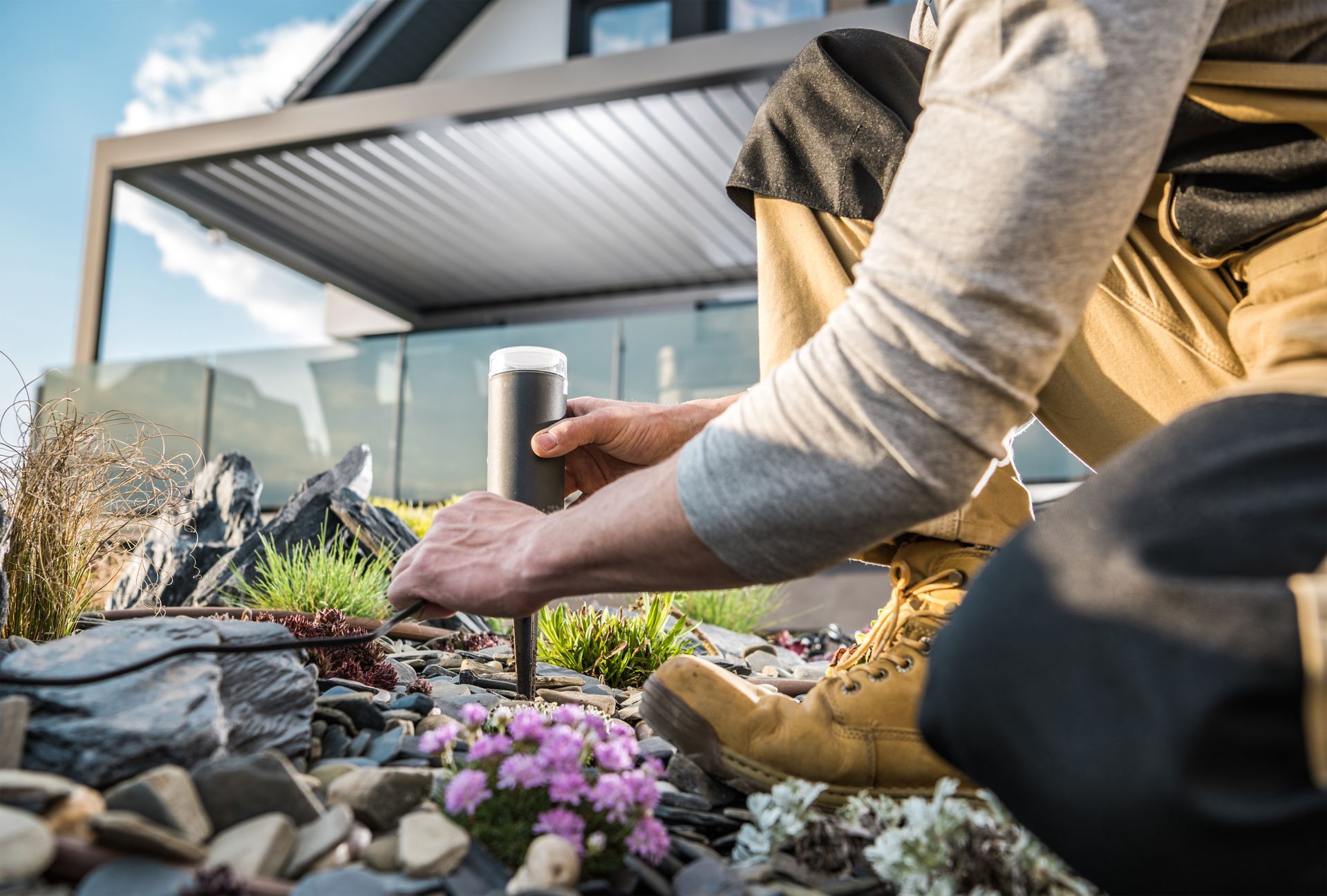 Person planting flowers in a garden bed beside a modern house under a patio roof.