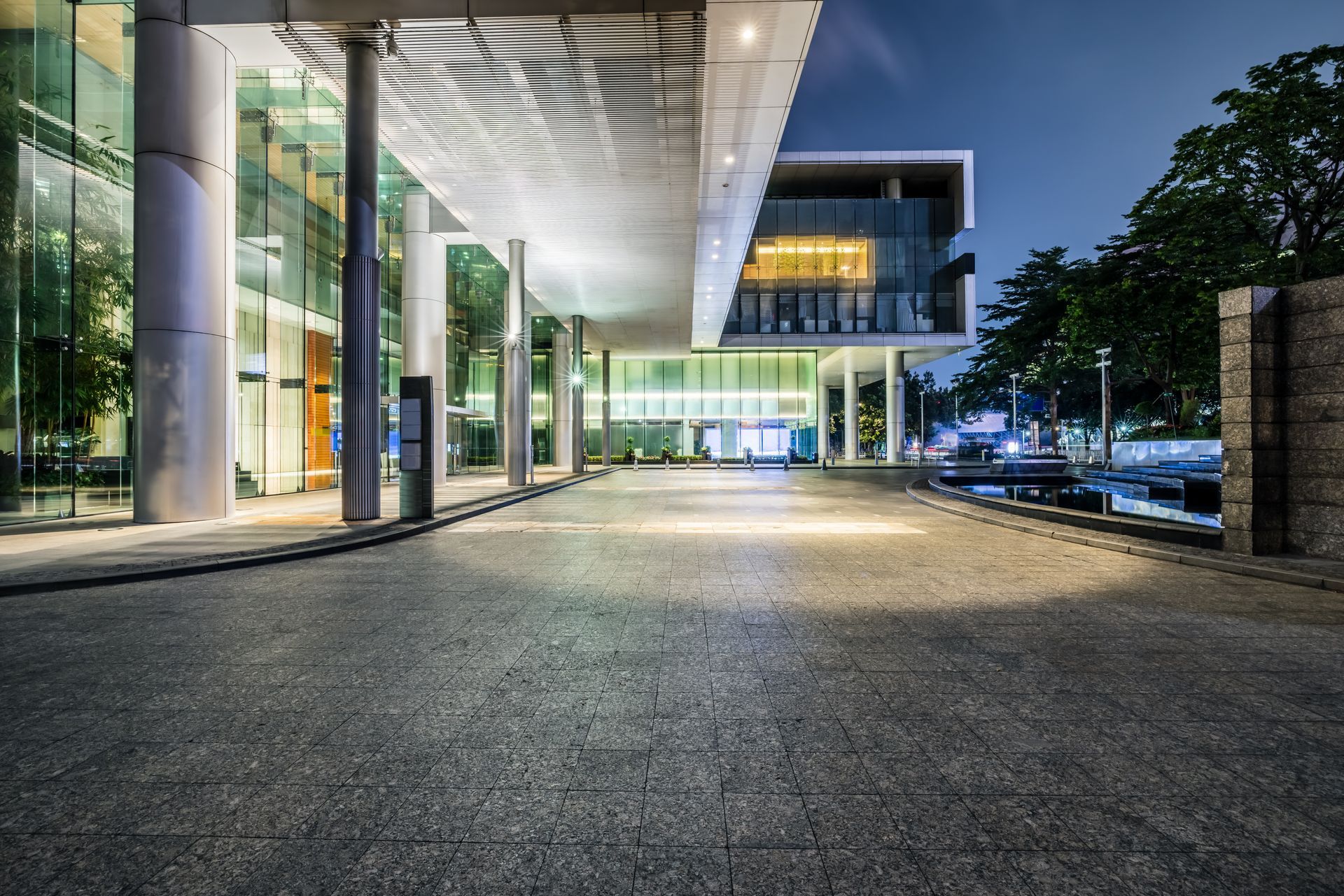 Modern glass building entrance at night with bright lights and an empty paved walkway.