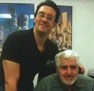Two men posing indoors, one standing behind the other, both smiling near cityscape wall art.