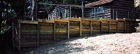 Wooden retaining wall in front of a log cabin. Sandy ground in the foreground.