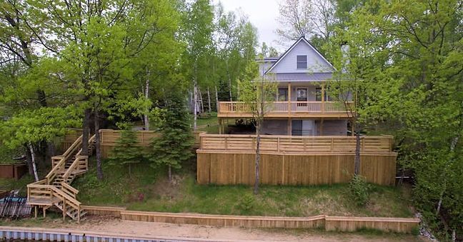 A two-story lakeside house with a wooden deck, fence, and stairs, surrounded by green trees.