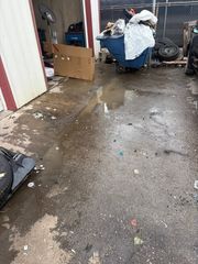 Trash-strewn alley with a blue bin, cardboard, and a puddle on wet pavement