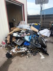 Overflowing trash pile beside an open garage in a driveway, with scattered debris and a black trash bag.