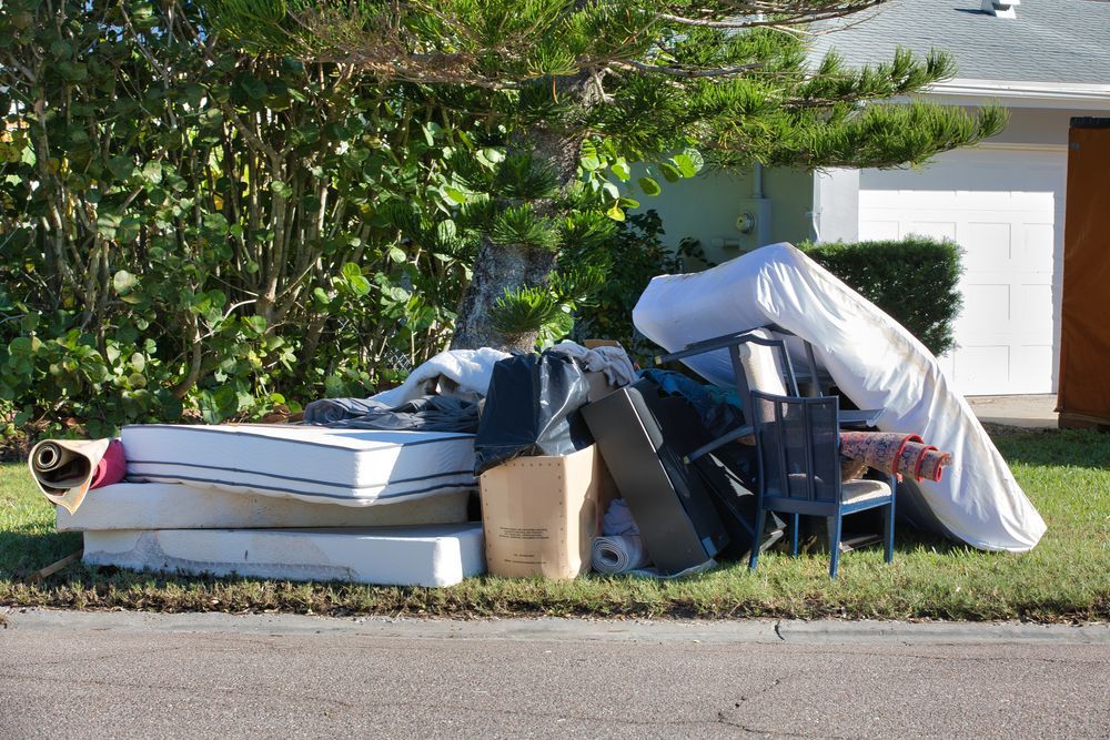 Piled curbside trash and covered furniture left on a residential street by a white garage door