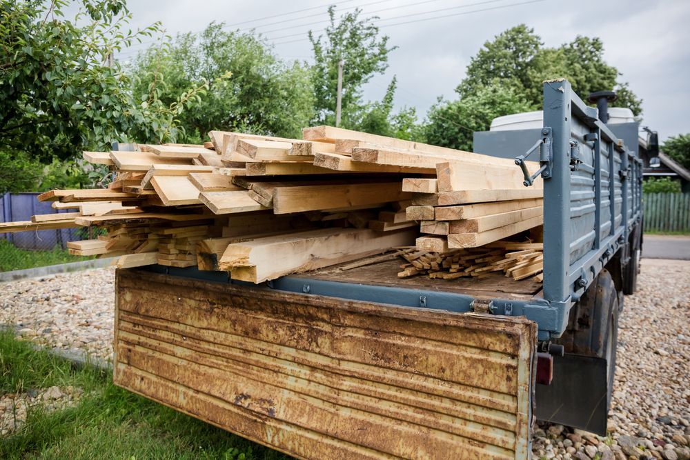 Pile of lumber on a flatbed trailer parked outdoors on gravel.