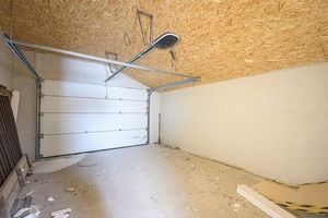 Empty garage with white roll-up door, unfinished OSB ceiling, and a bare concrete floor.