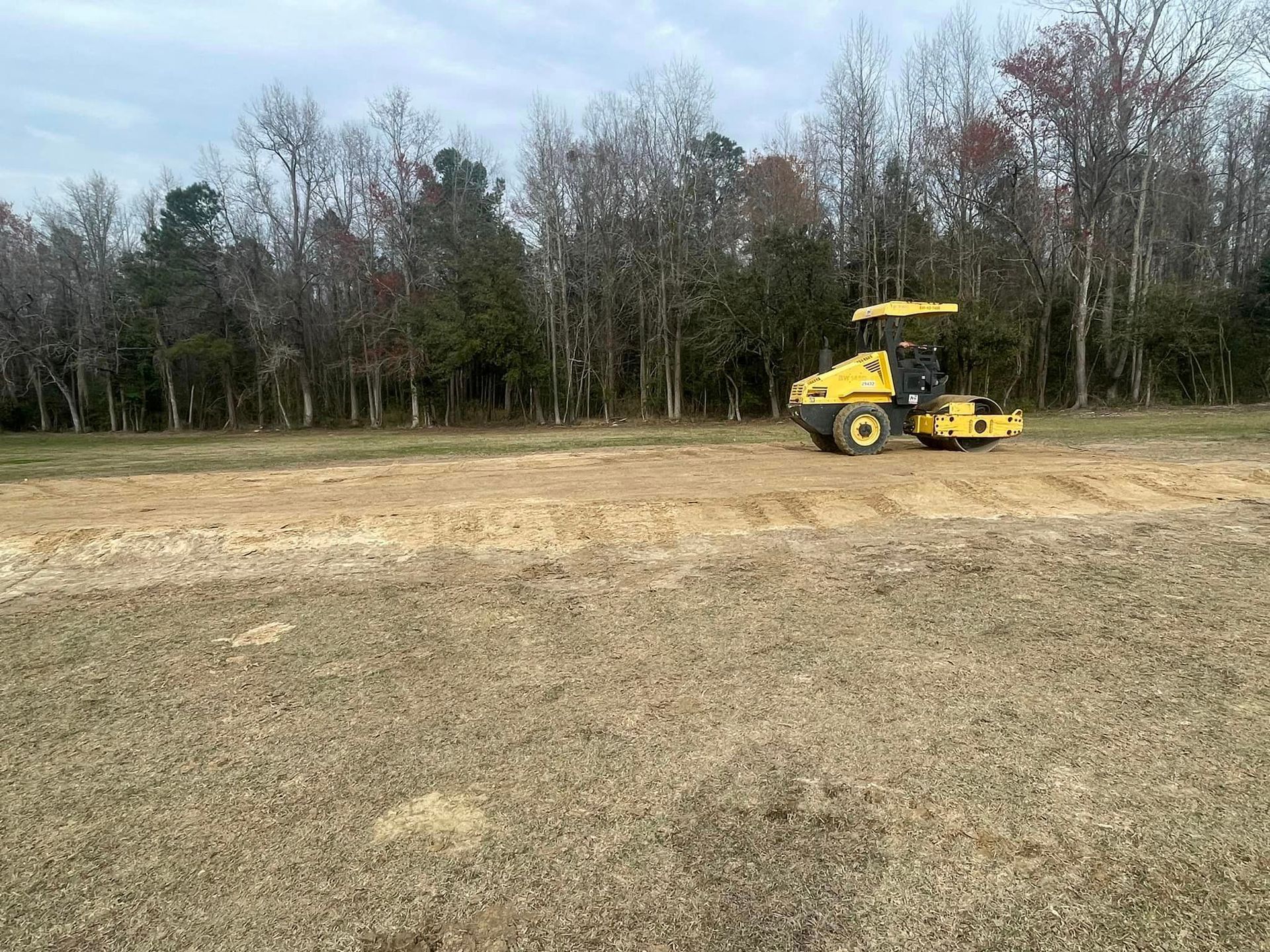 A yellow roller is rolling dirt in a field with trees in the background.