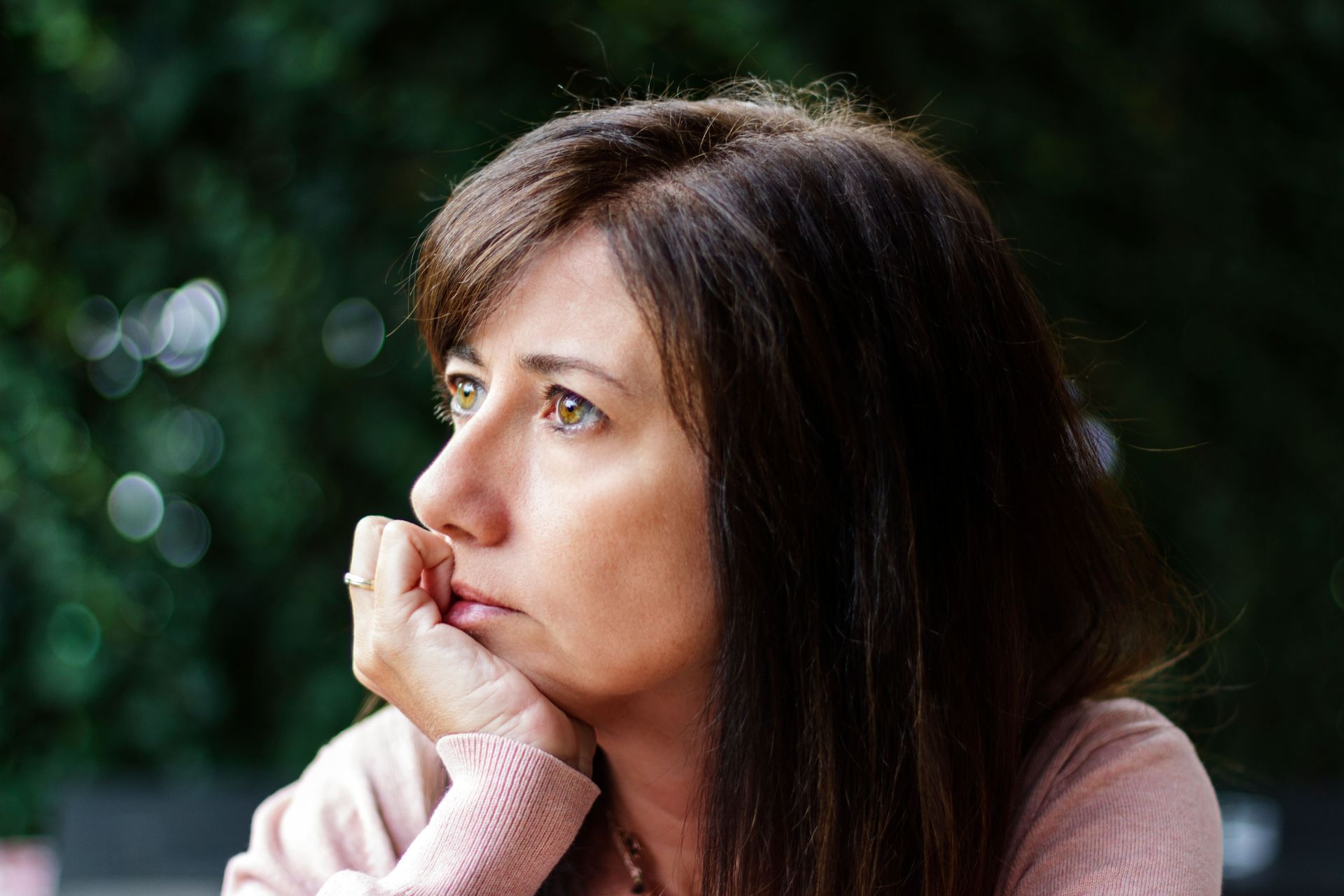 Woman with brown hair resting chin on hand, looking off to the side with a pensive expression; blurred green background.