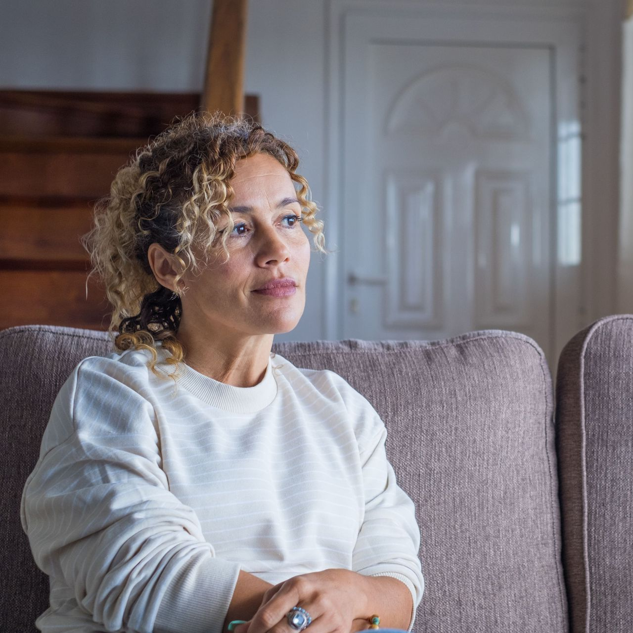 Woman sitting on a couch, looking to the side with a pensive expression, light-filled room.