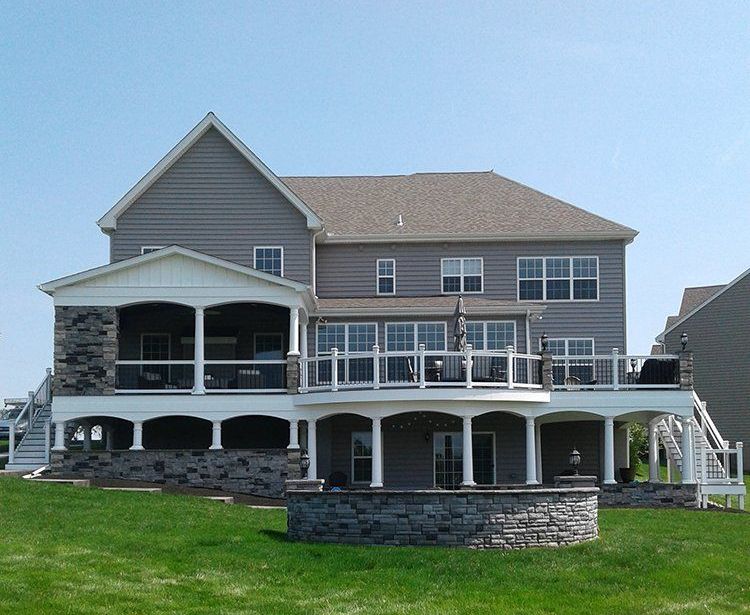 Large gray suburban house with a wraparound porch and stone foundation on a grassy lawn