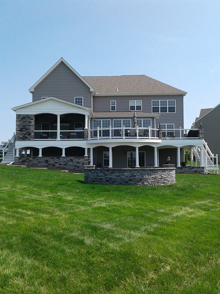 Large gray beach house with wraparound porch on a grassy hillside under a clear sky
