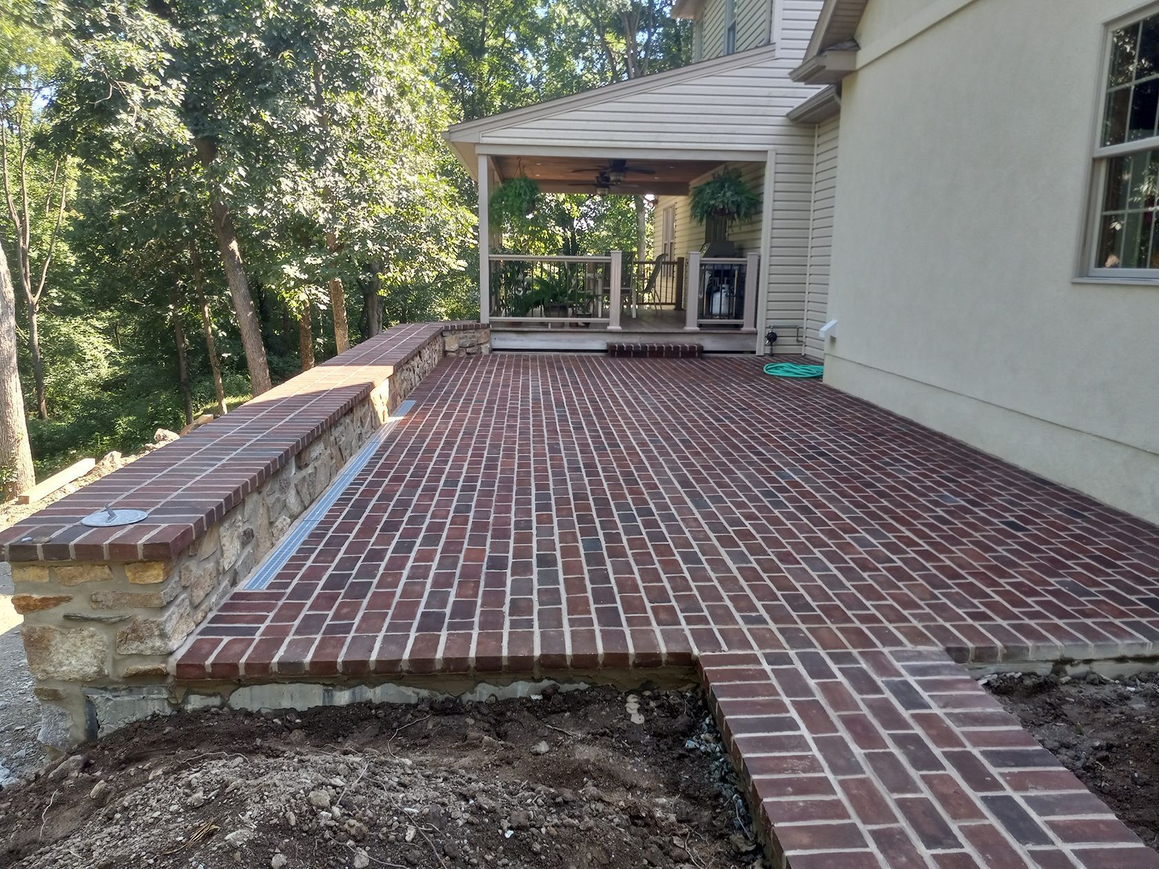 Brick patio beside a white house, with a stone planter wall and wooded backyard beyond.