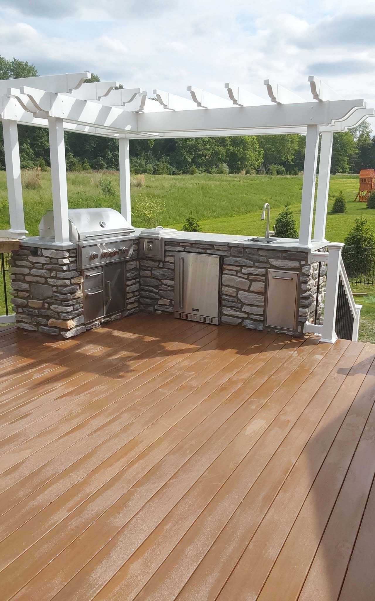 Wooden deck with a white pergola and stone outdoor kitchen overlooking a green lawn