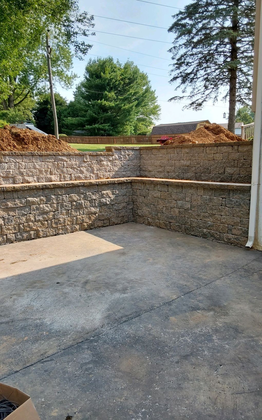 Stone retaining wall and concrete patio under construction, with trees and soil mounds in the background.