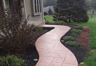 Curved pink walkway beside a house, bordered by mulch and shrubs in a landscaped yard.