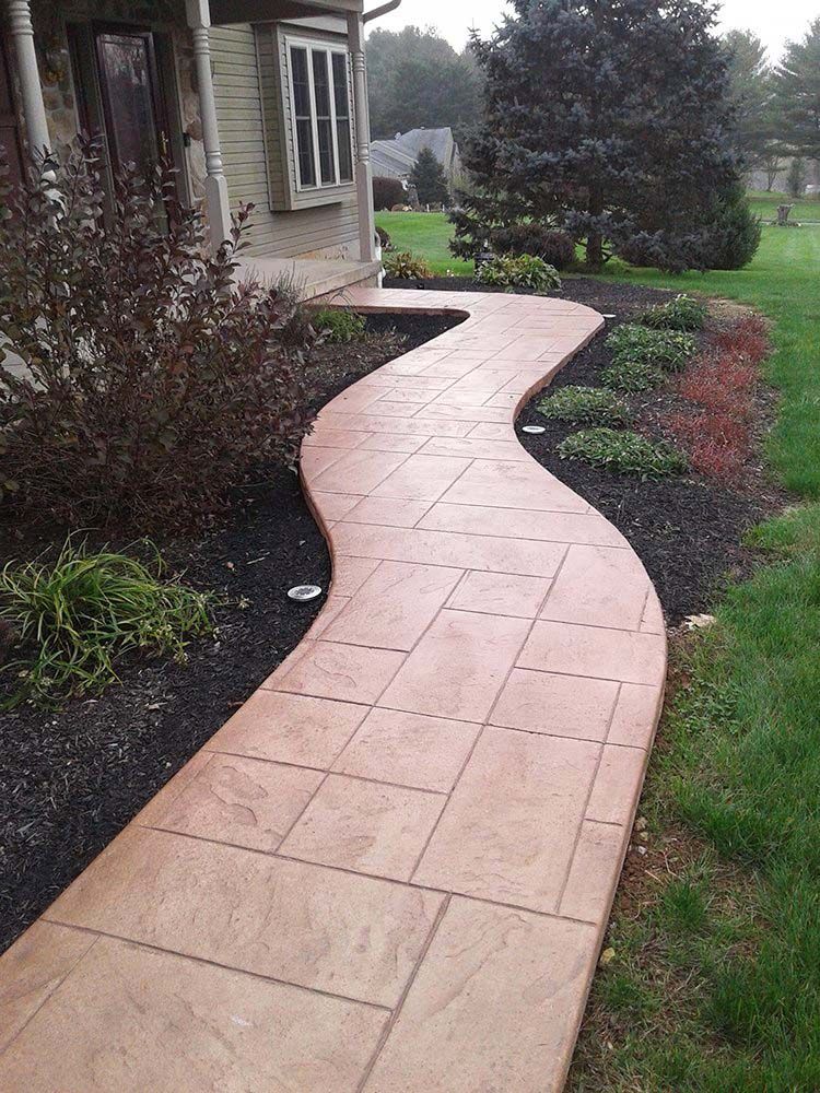 Curving pink-tiled garden walkway beside a house, bordered by shrubs and green lawn.