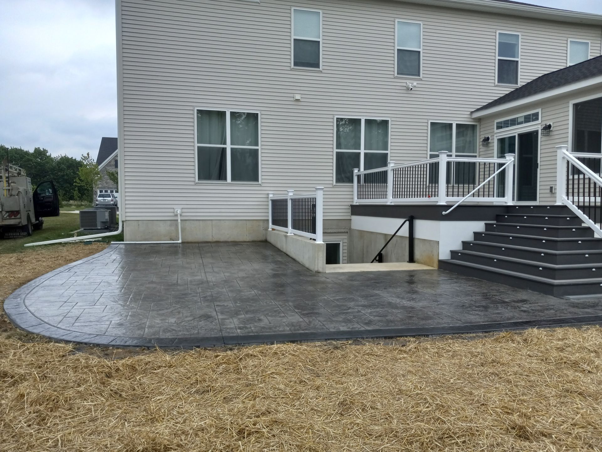 Backyard patio beside a gray house, with steps, railing, and a gravel yard.