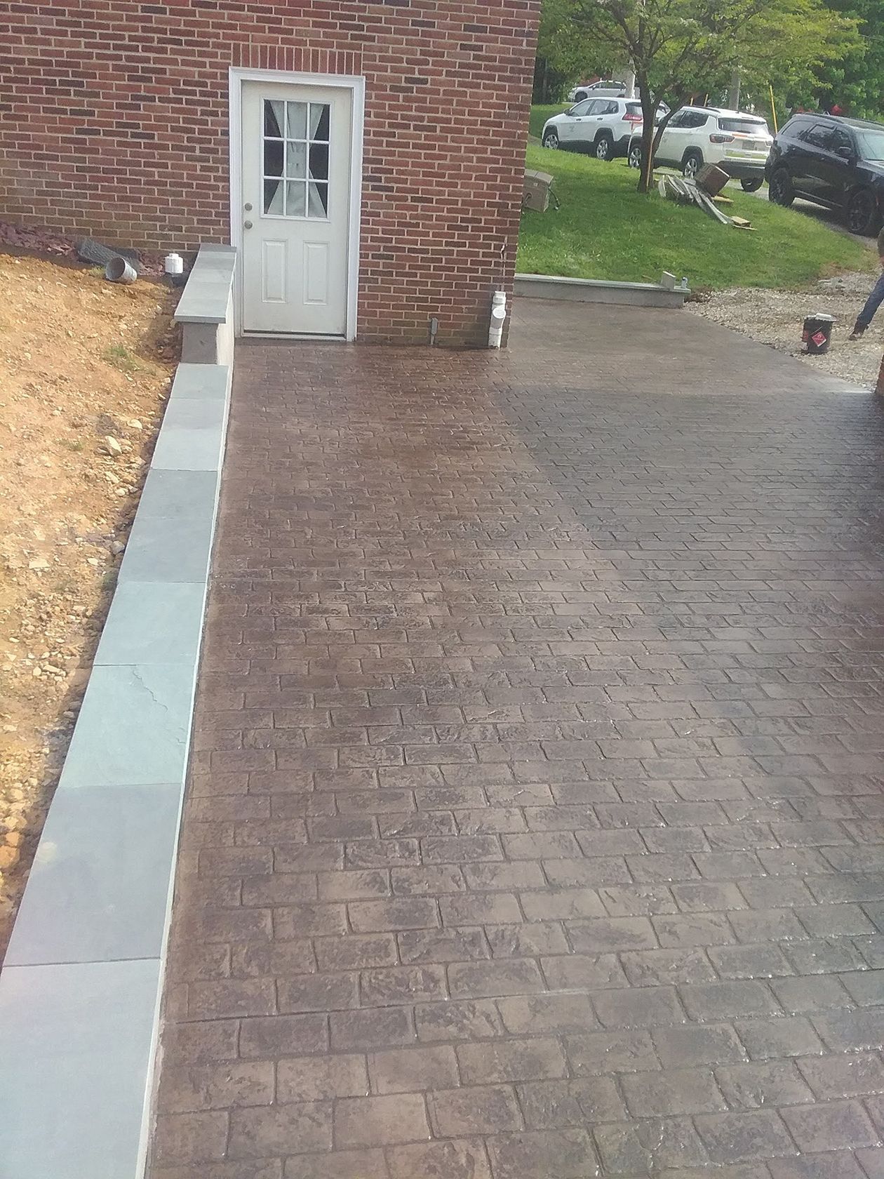 Brick driveway beside a house, with a gray door and steps leading to a paved parking area.