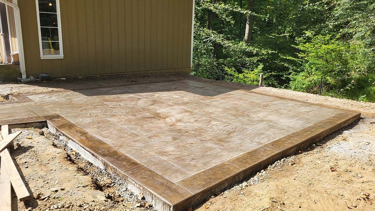 Concrete patio slab beside a house, surrounded by gravel and dirt, with a wooden border