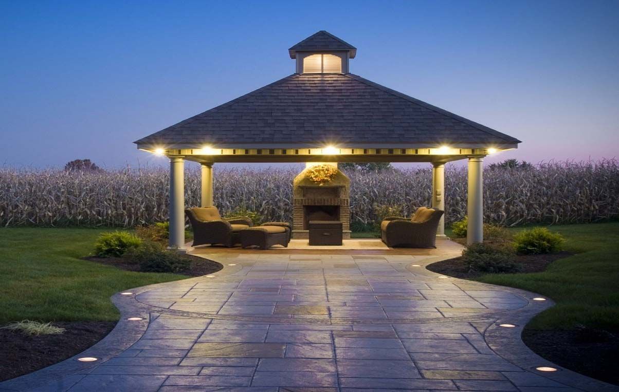 Lit gazebo at dusk with stone path leading to it, set beside a field of tall grasses.