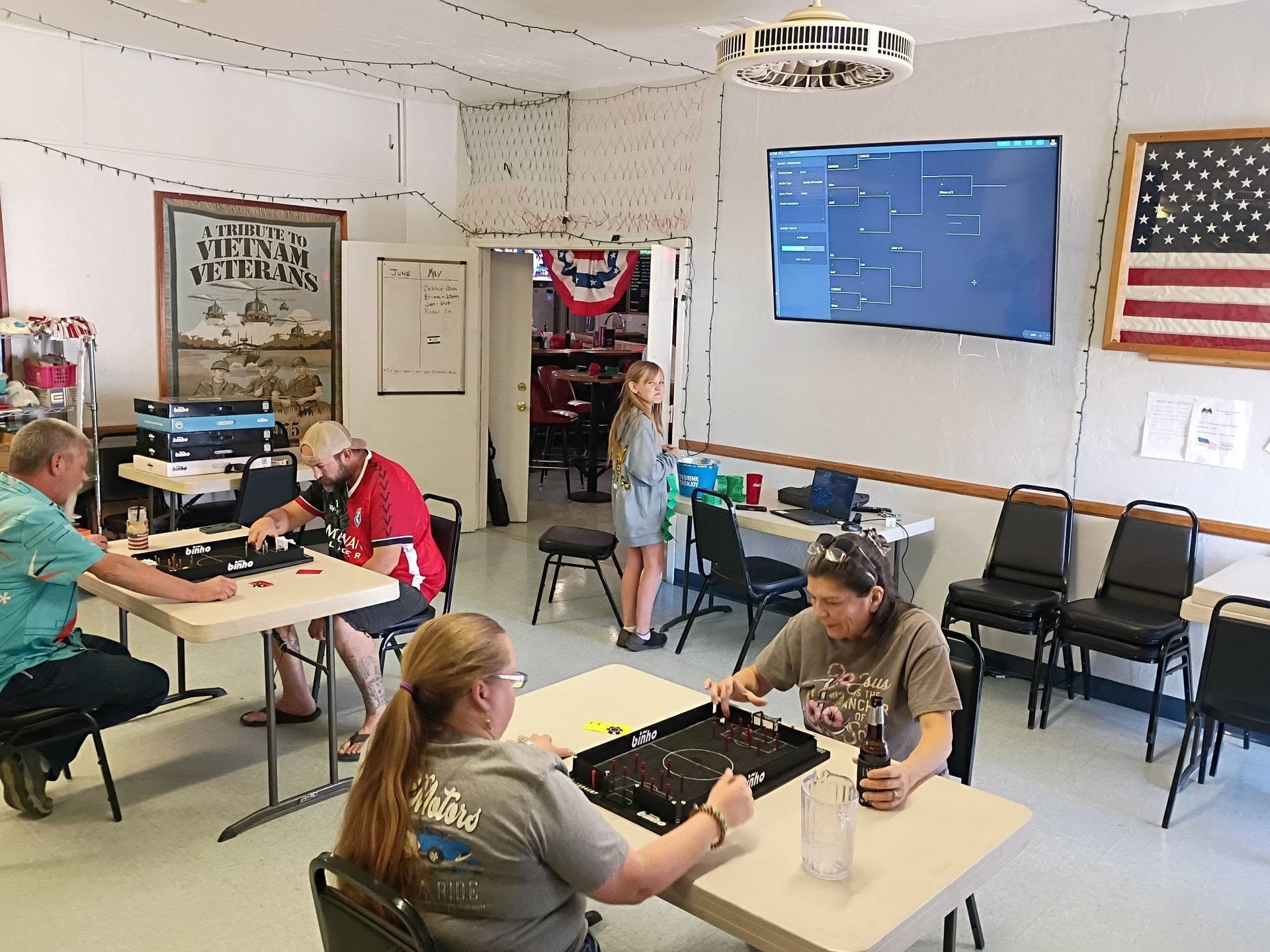 People seated at tables in a small meeting room with a TV on the wall and an American flag displayed.