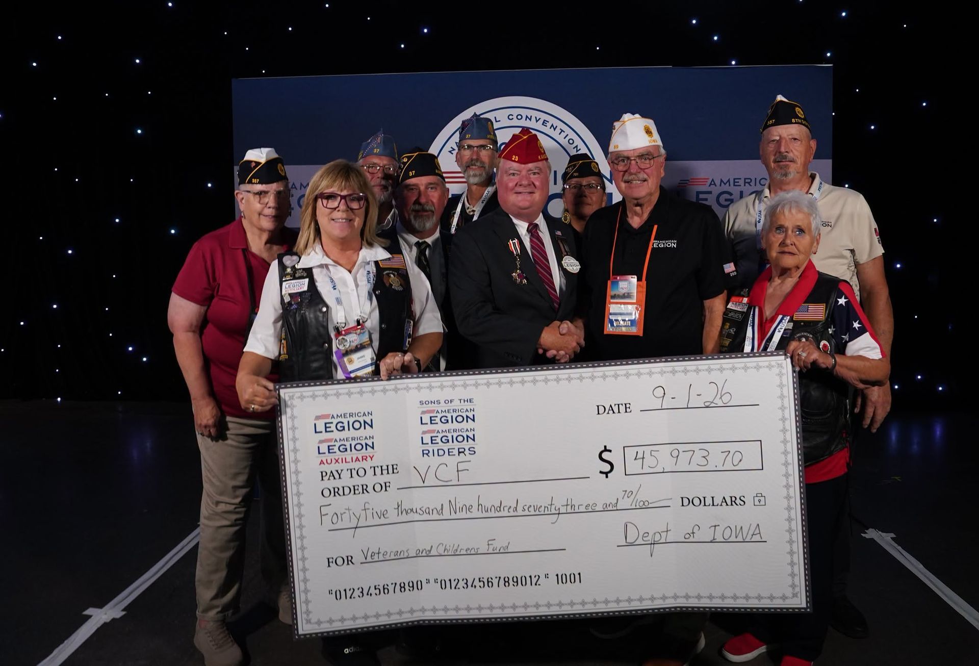 Group holding an oversized check on a stage with a starry backdrop, smiling in event attire.