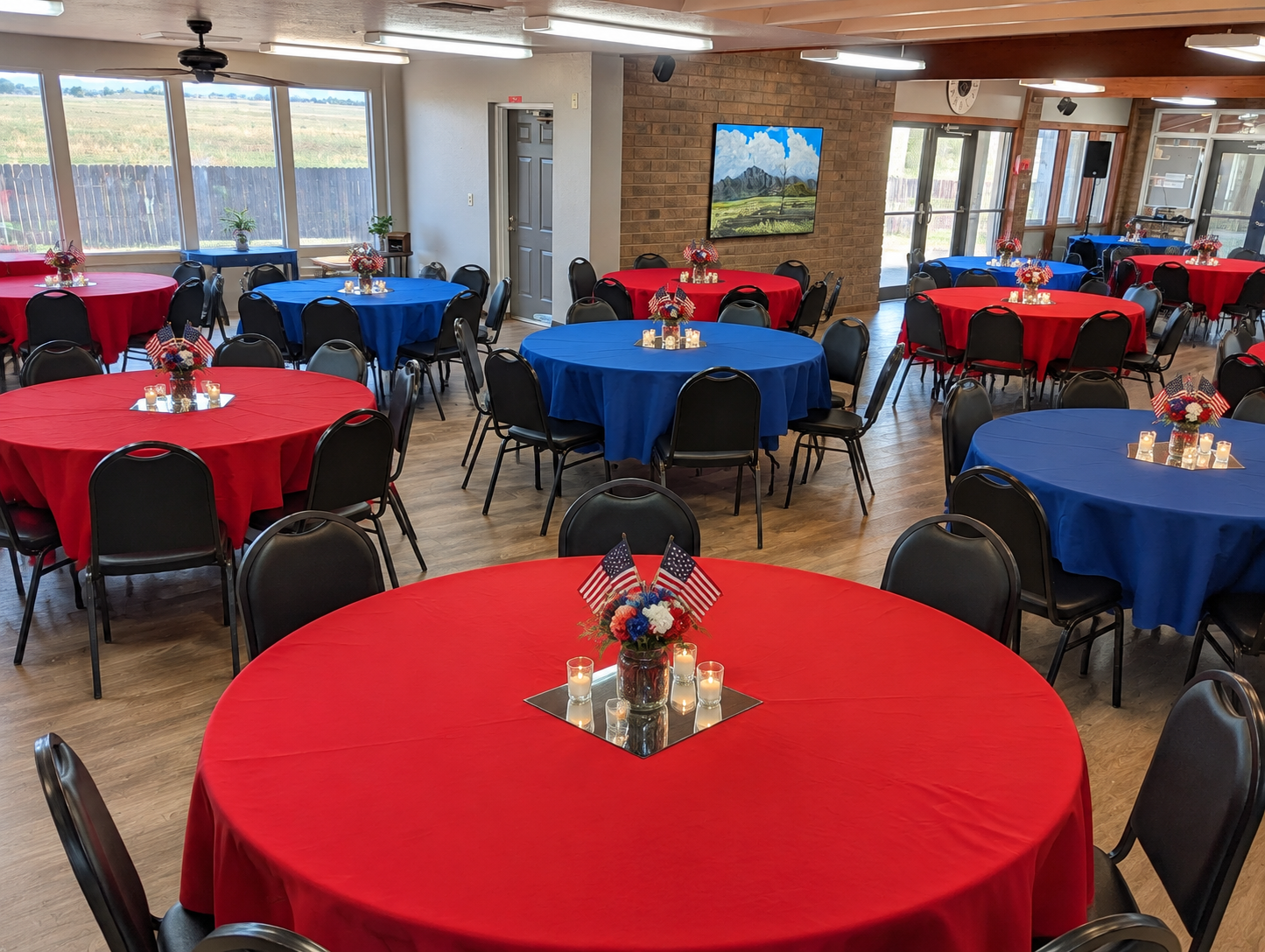 Empty banquet room with red and blue tablecloths, black chairs, and windows along the wall.