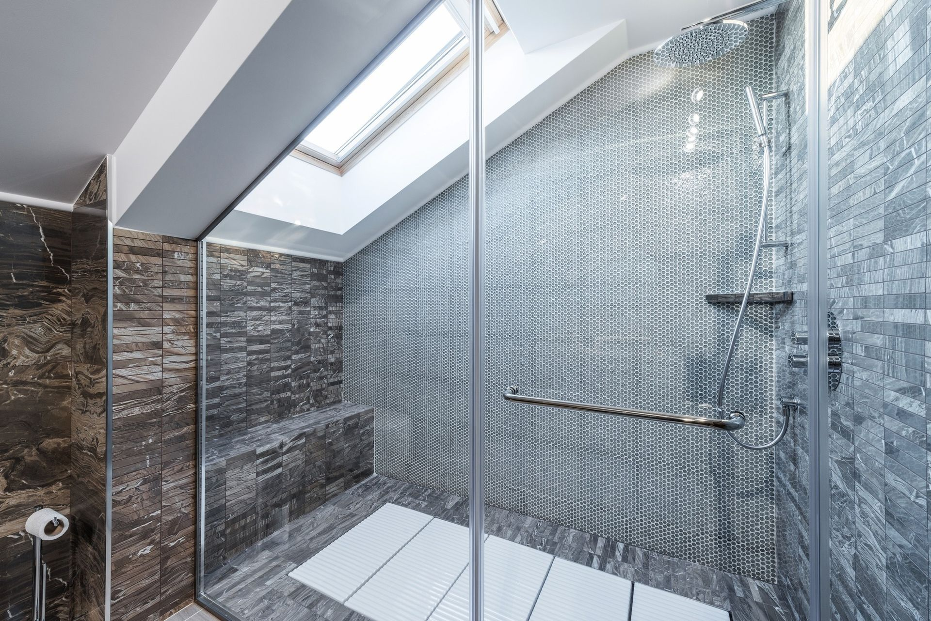 Shower with glass door, gray textured tile walls, white floor mat, and skylight.