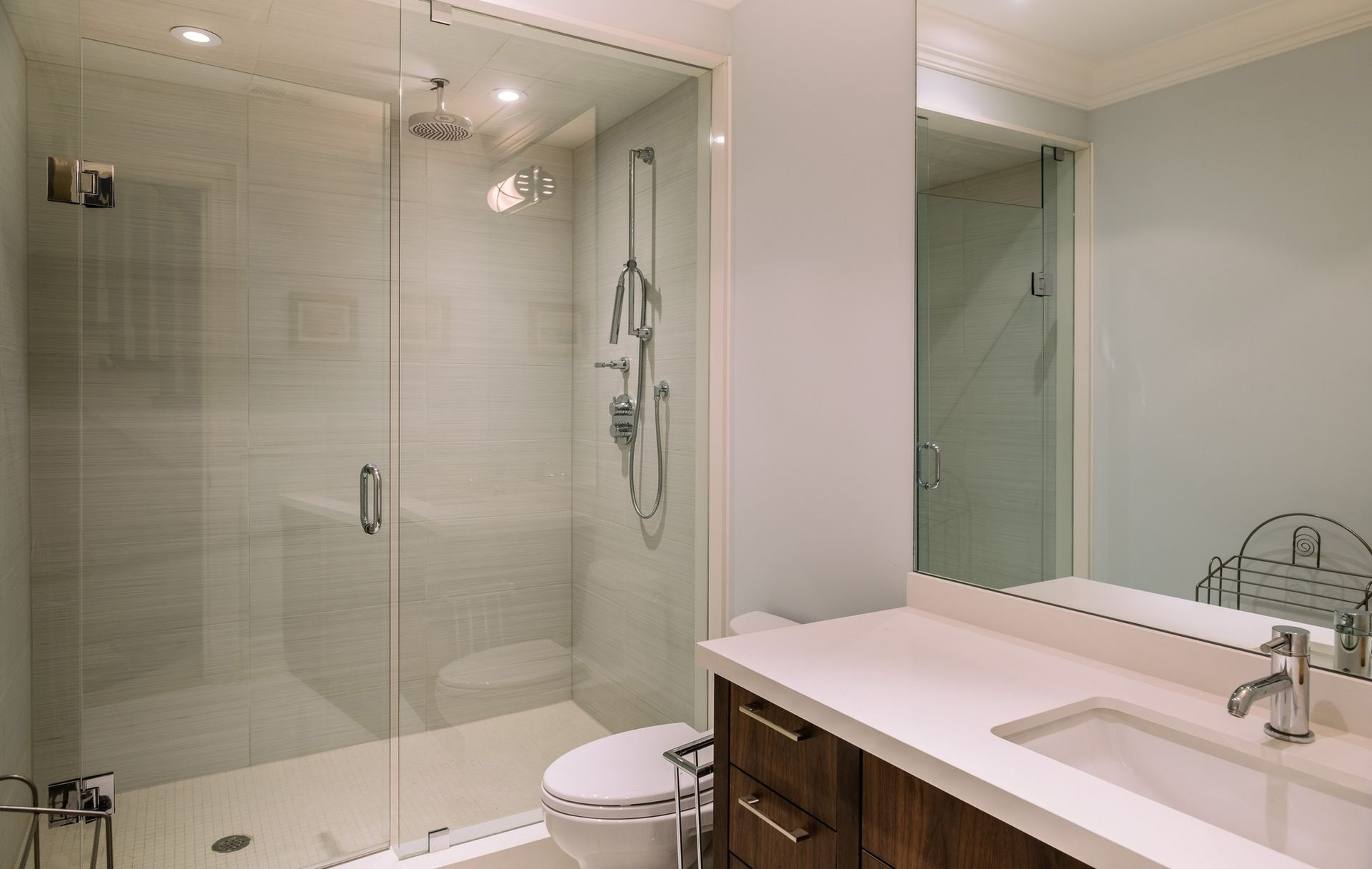 Modern bathroom with a glass shower, toilet, vanity, and mirror. Light blue walls and white countertop.