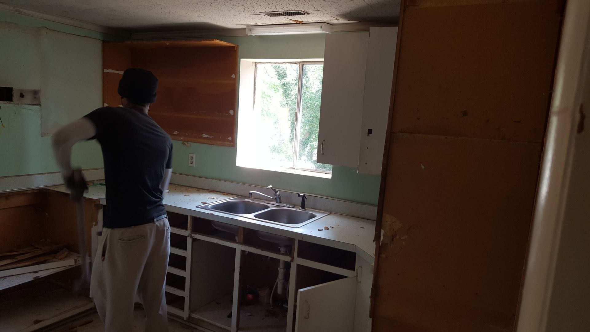 A person in a black t-shirt works on renovating a kitchen, removing cabinets beneath a window above a dual kitchen sink.