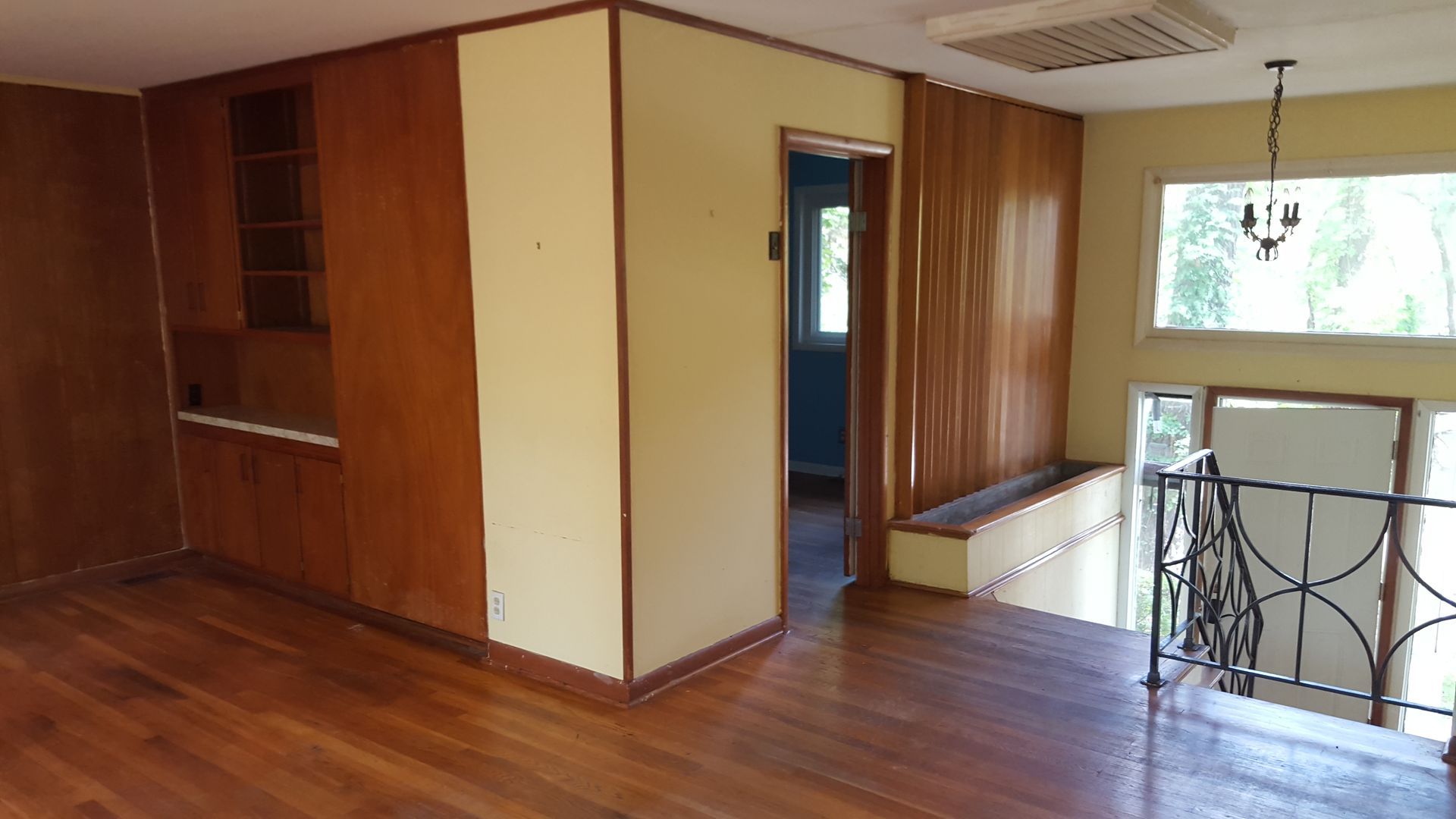 A split-level interior with wood-paneled walls, hardwood floors, a yellow partition wall, and a metal-railed staircase.