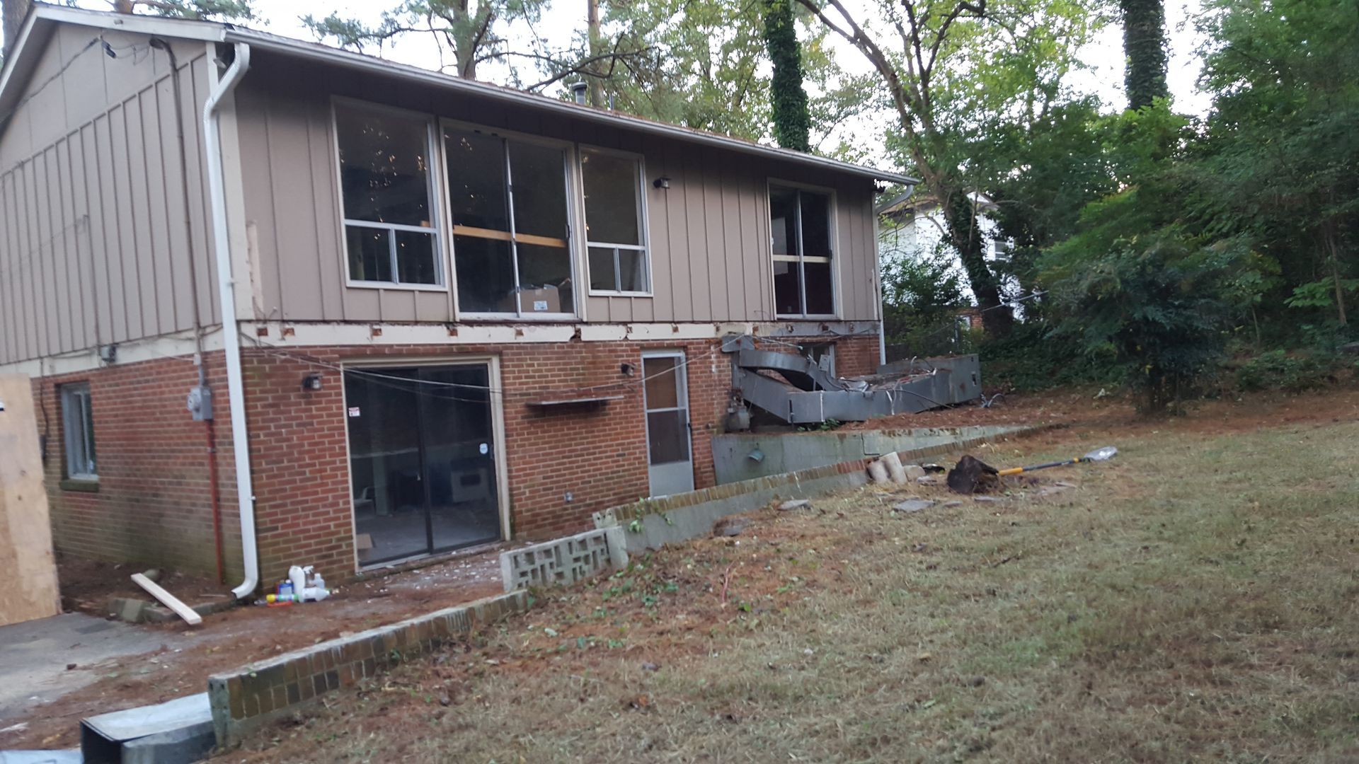 A two-story house under construction with a brick lower level and wood-paneled upper level, surrounded by wooded yard.