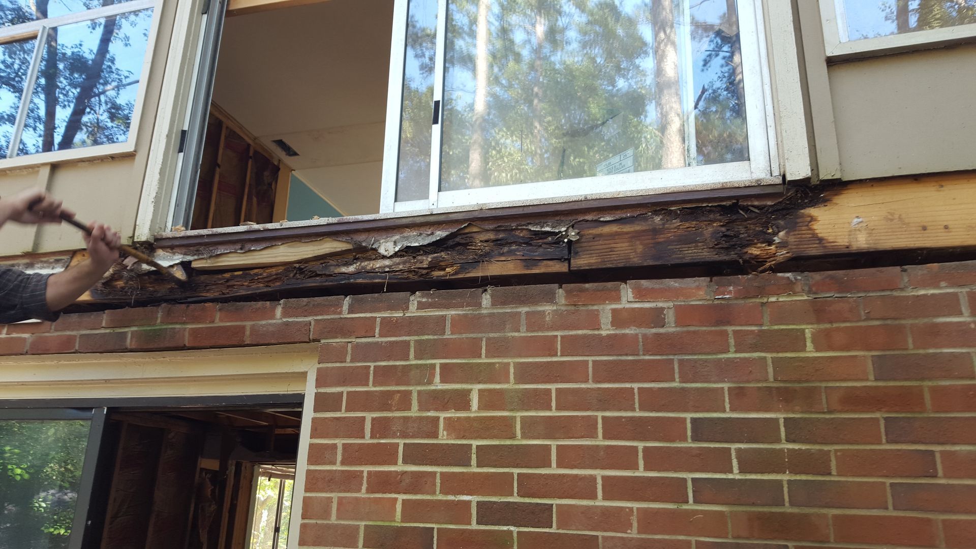 A person uses a pry bar to remove rotted wood from a deck ledger board attached to a brick house below a sliding door.