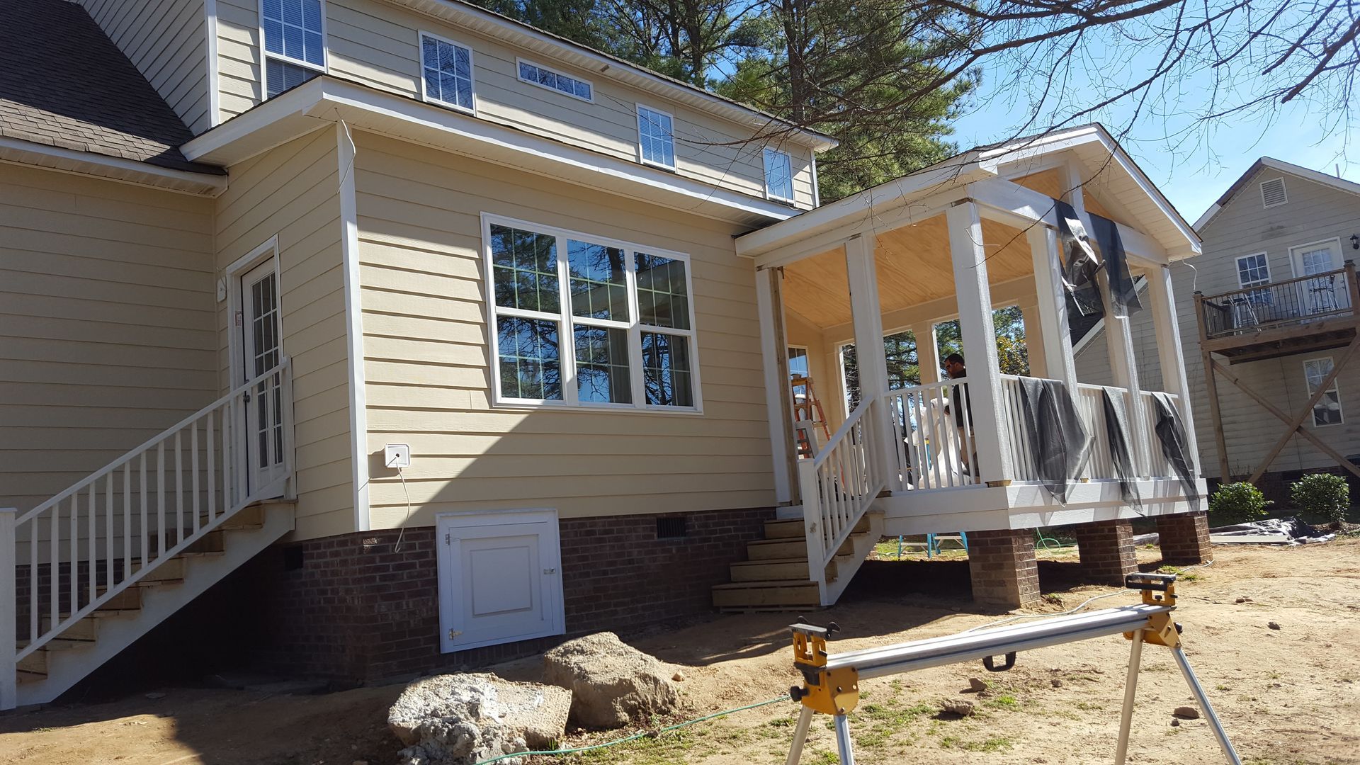 Exterior view of a light yellow house under construction with a back deck, stone foundation, and a portable work stand.