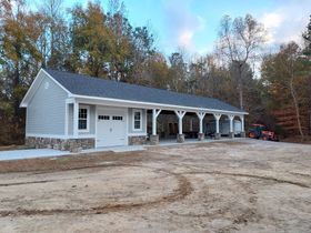 New gray house with stone accents and covered porch in a wooded setting with a dirt driveway