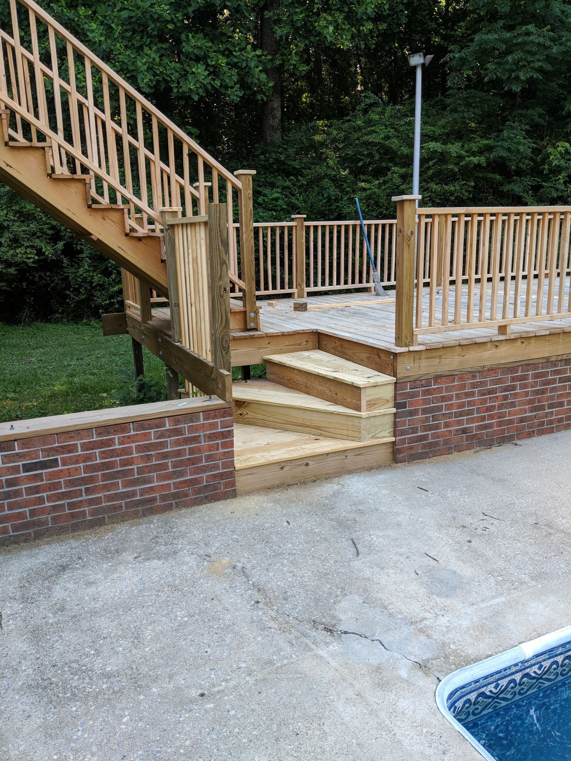 A wooden deck and staircase with natural-finish balusters, situated next to a brick retaining wall and swimming pool.