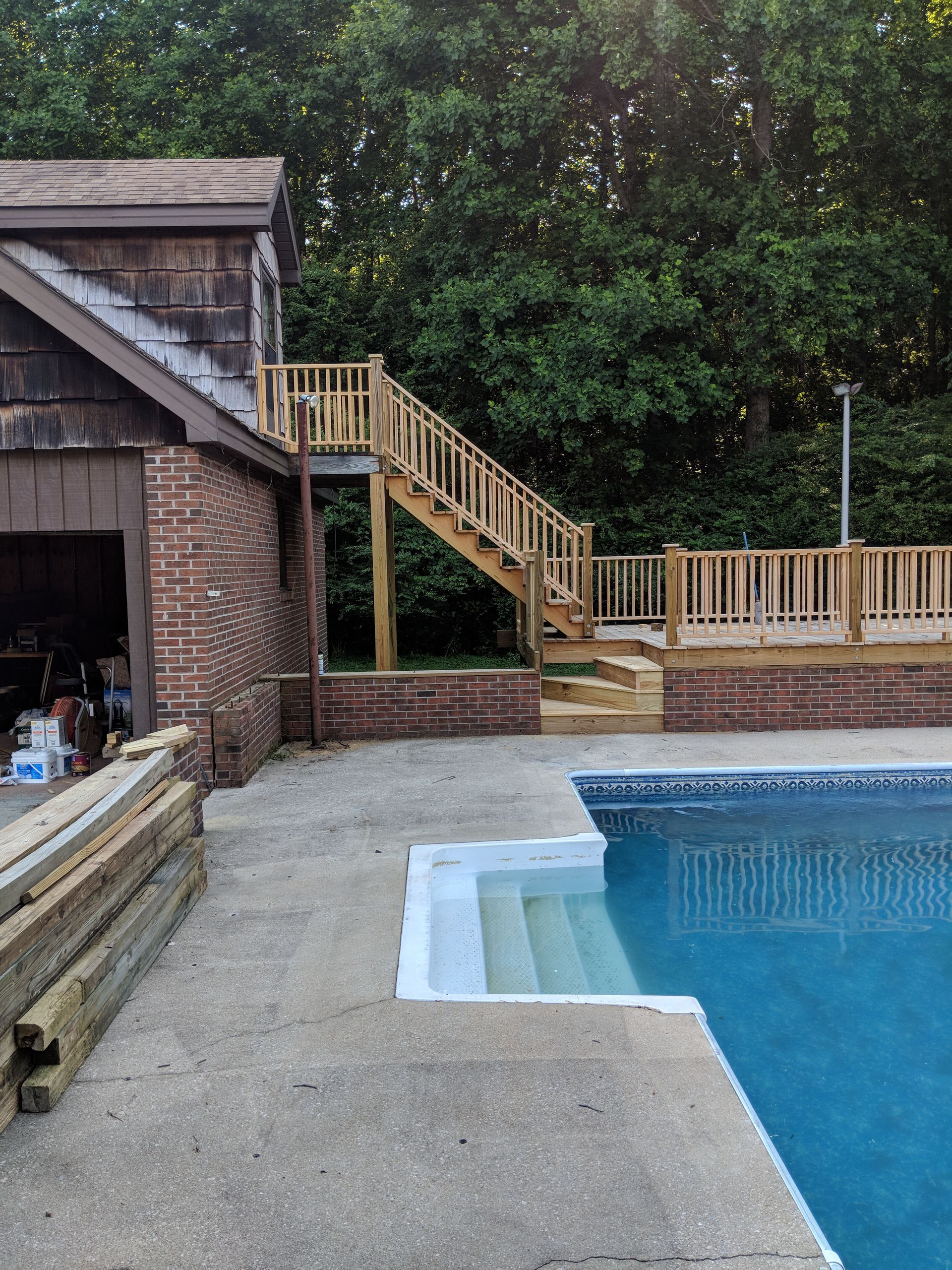 A swimming pool next to a brick building with a newly constructed wooden deck, staircase, and stone retaining wall.