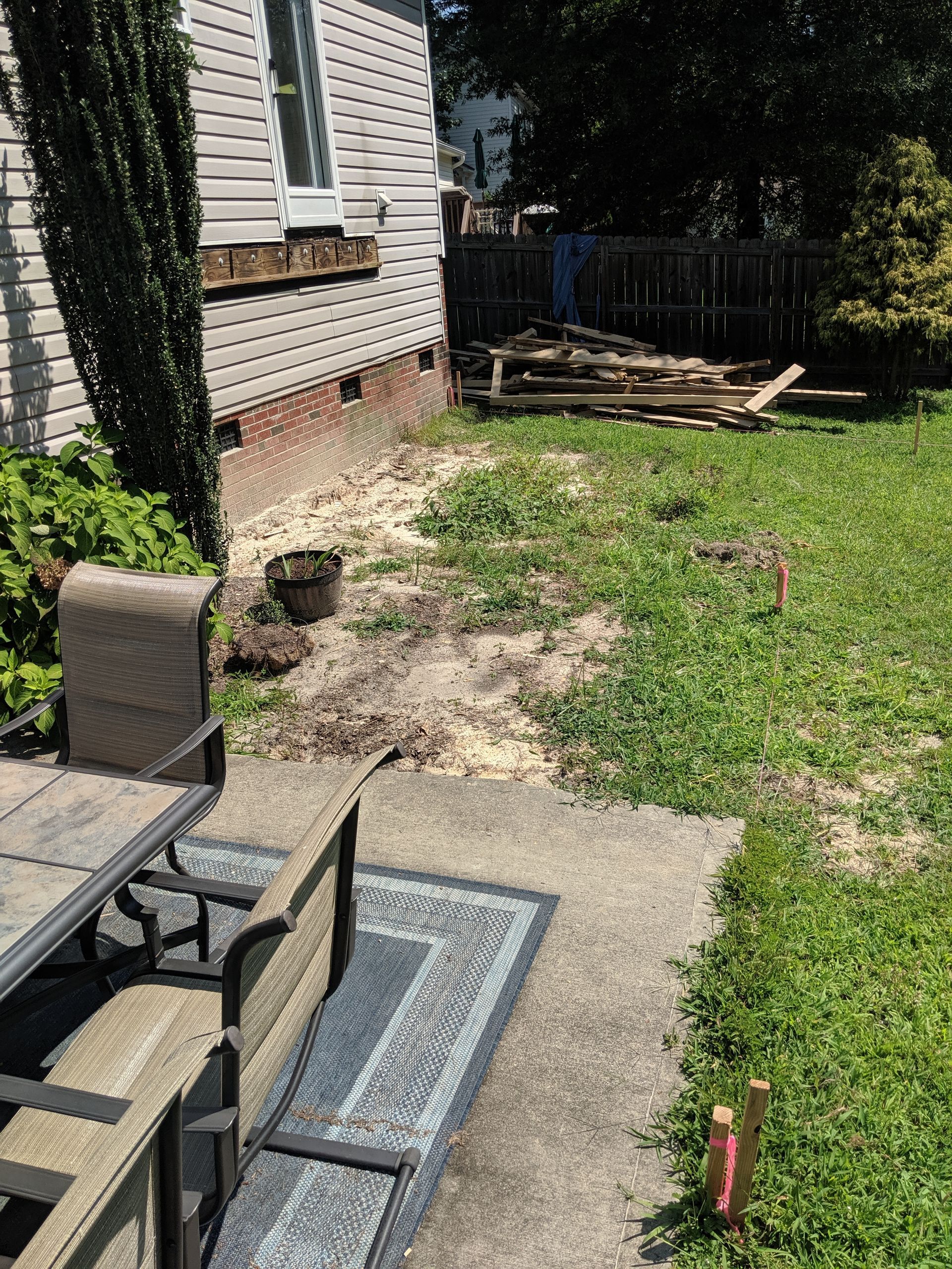 A patio set sits on a concrete slab next to a house with beige siding, leading to an area with sparse grass and debris.