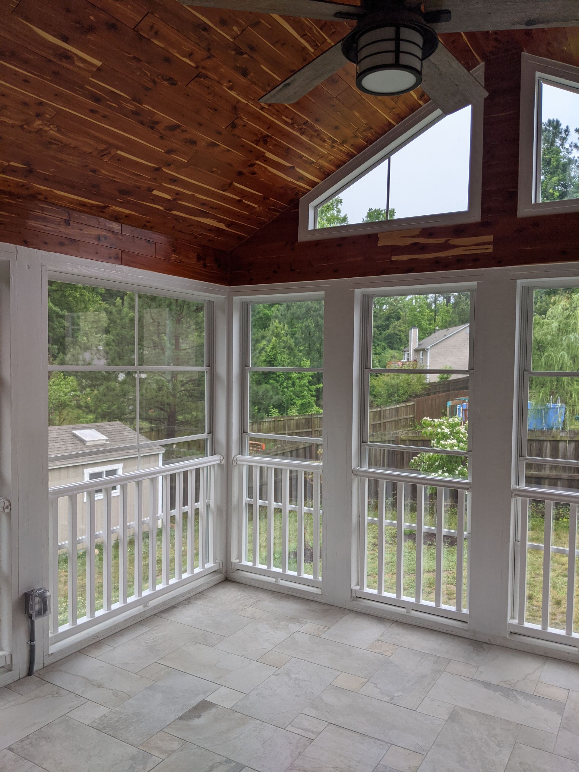 A sunroom with a wood-paneled vaulted ceiling, large screened windows, white railings, and light-colored stone flooring.