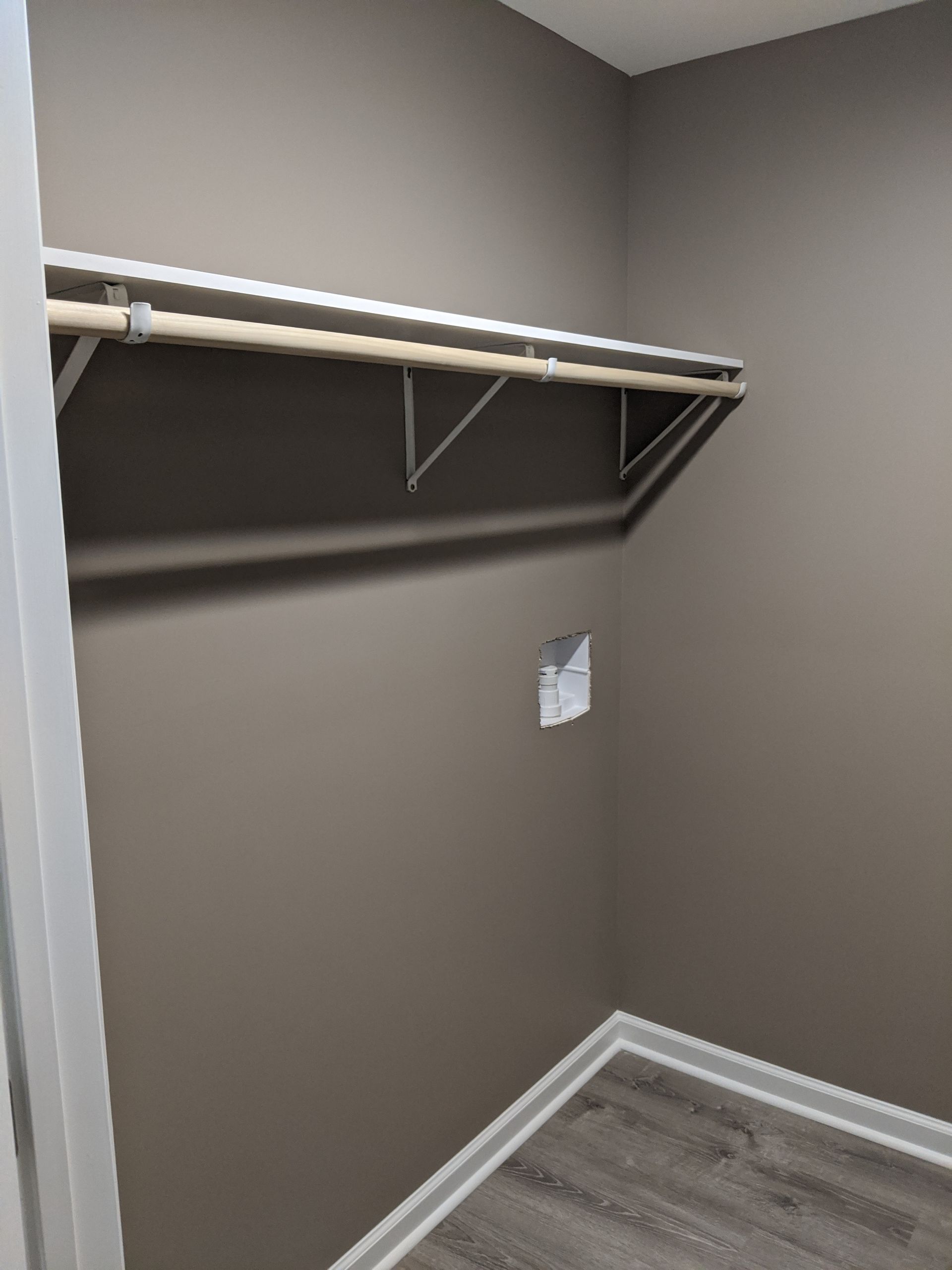 An empty closet with taupe walls, gray wood-look flooring, a white shelf, and a wooden clothes rod.