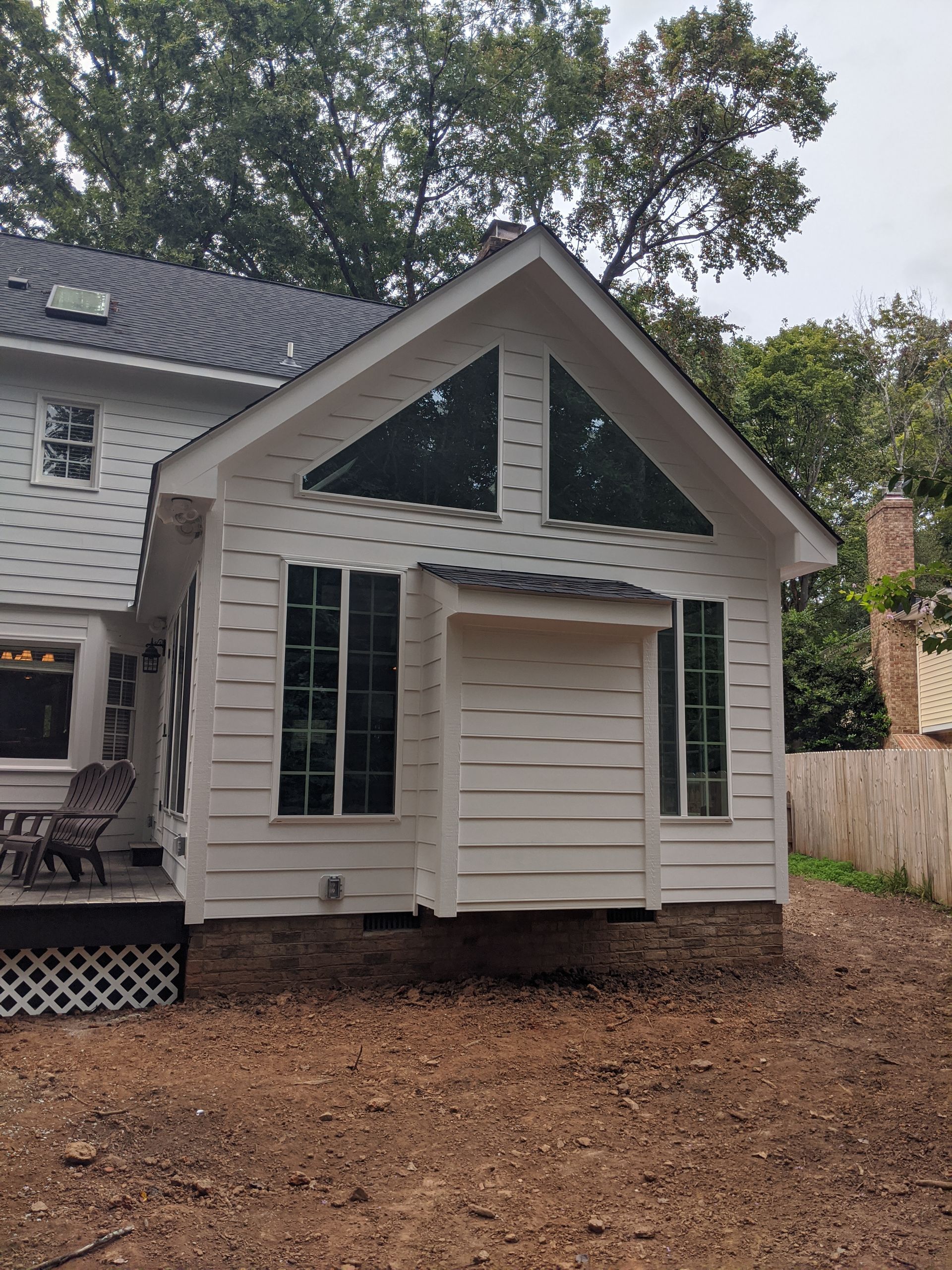 A white house extension with a gabled roof, large windows, and a rectangular bump-out, set in a yard with trees.
