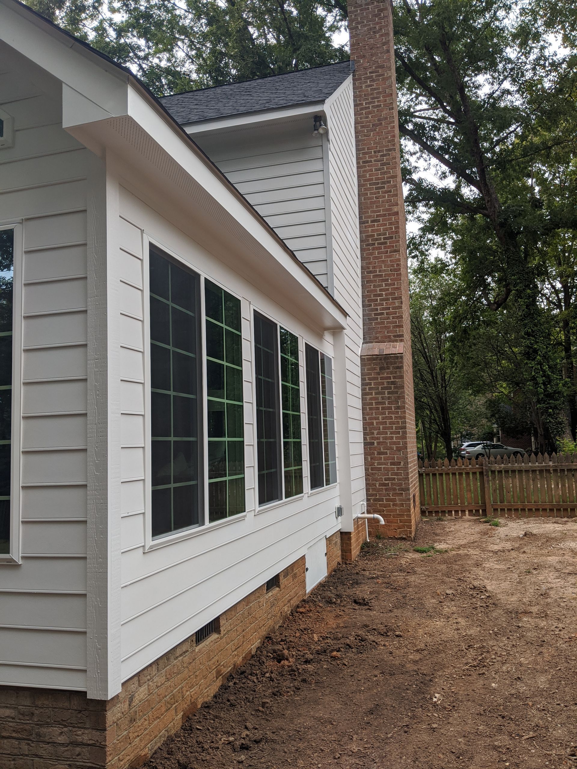 Side view of a white home with siding, dark-screened windows, a brick chimney, and an unfinished yard.