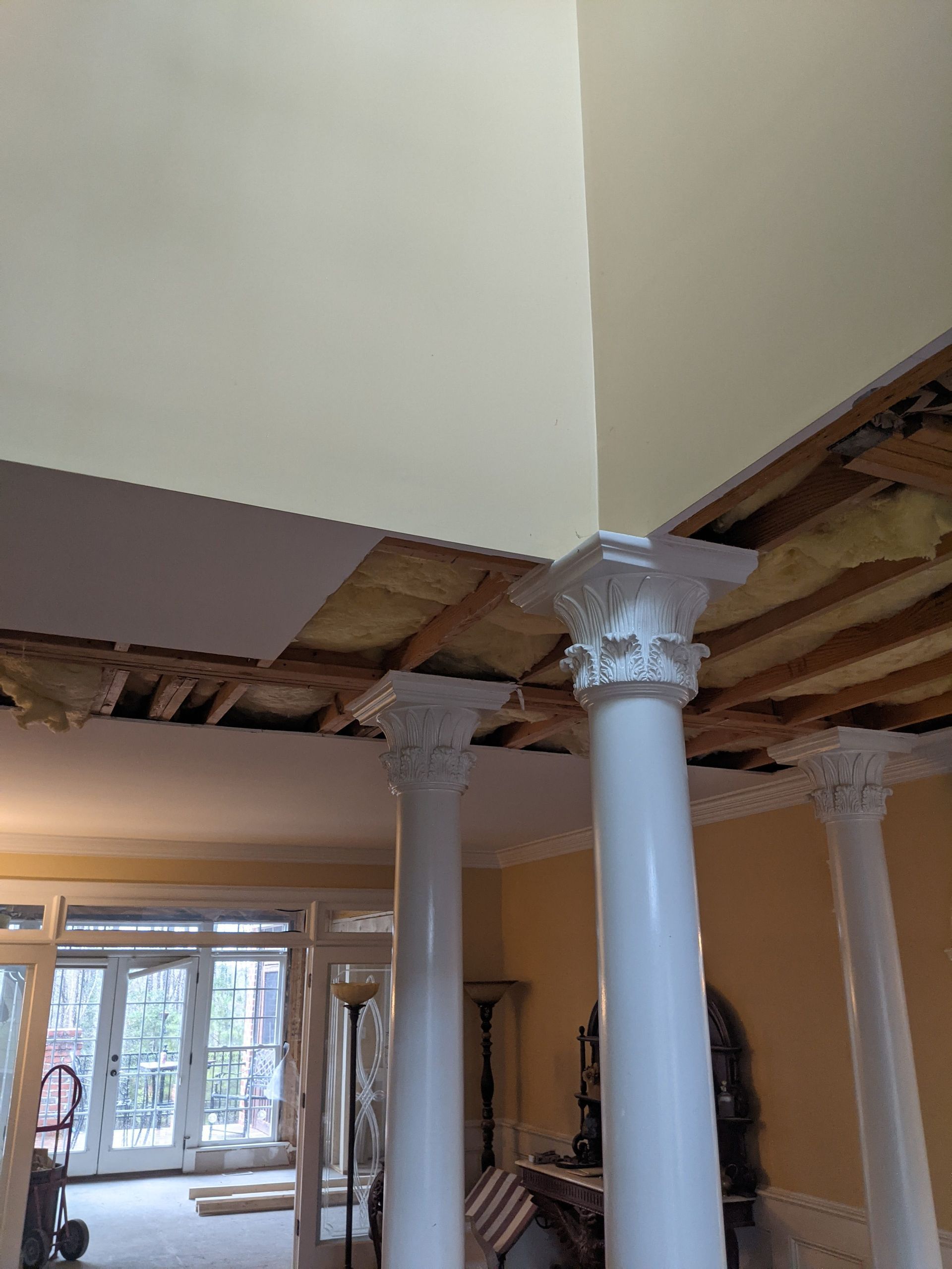 Interior view showing exposed ceiling joists and insulation above white Corinthian columns in a room under renovation.