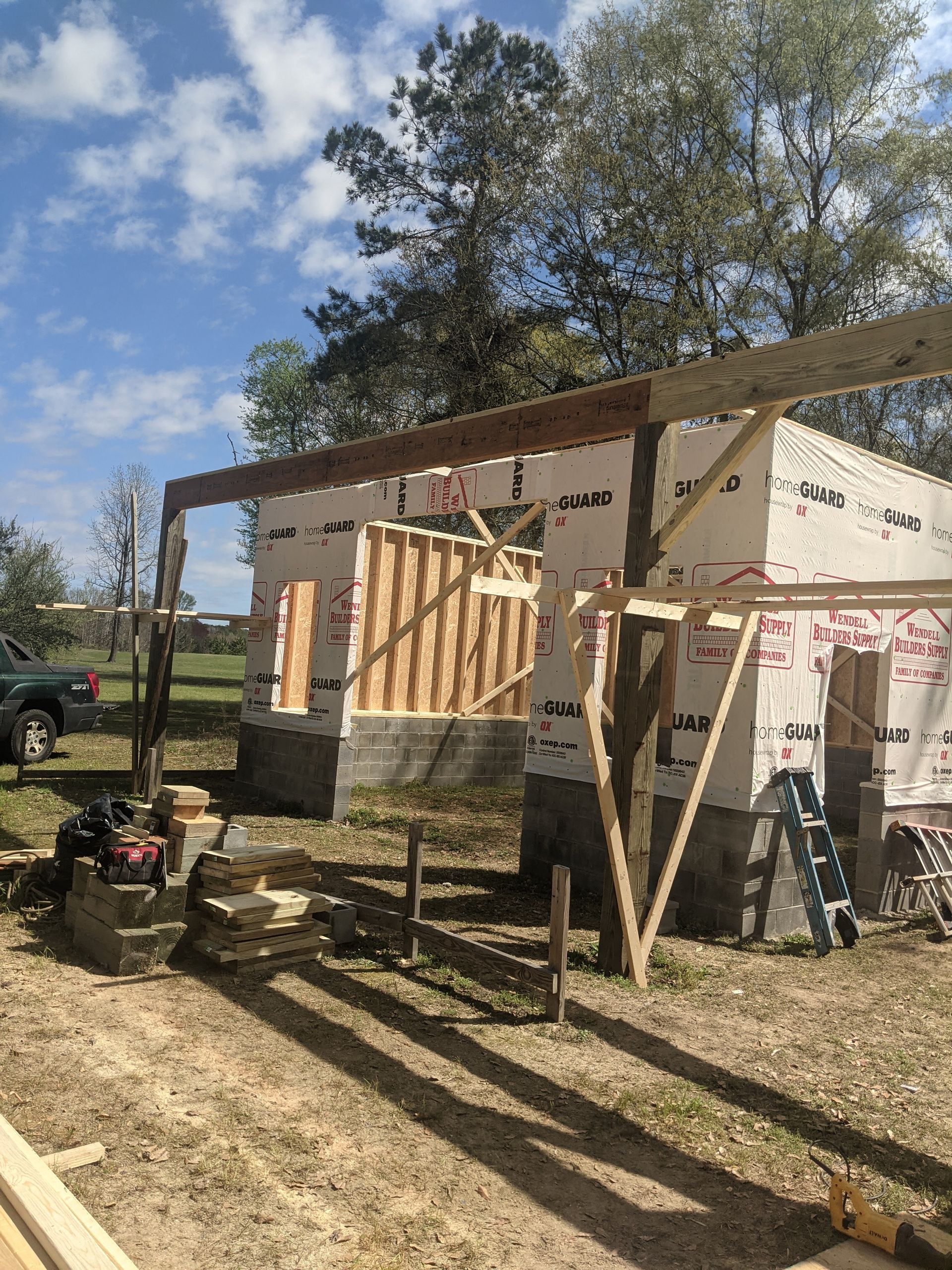 A wooden building frame under construction outdoors on a grassy lot, featuring a large overhanging support beam.