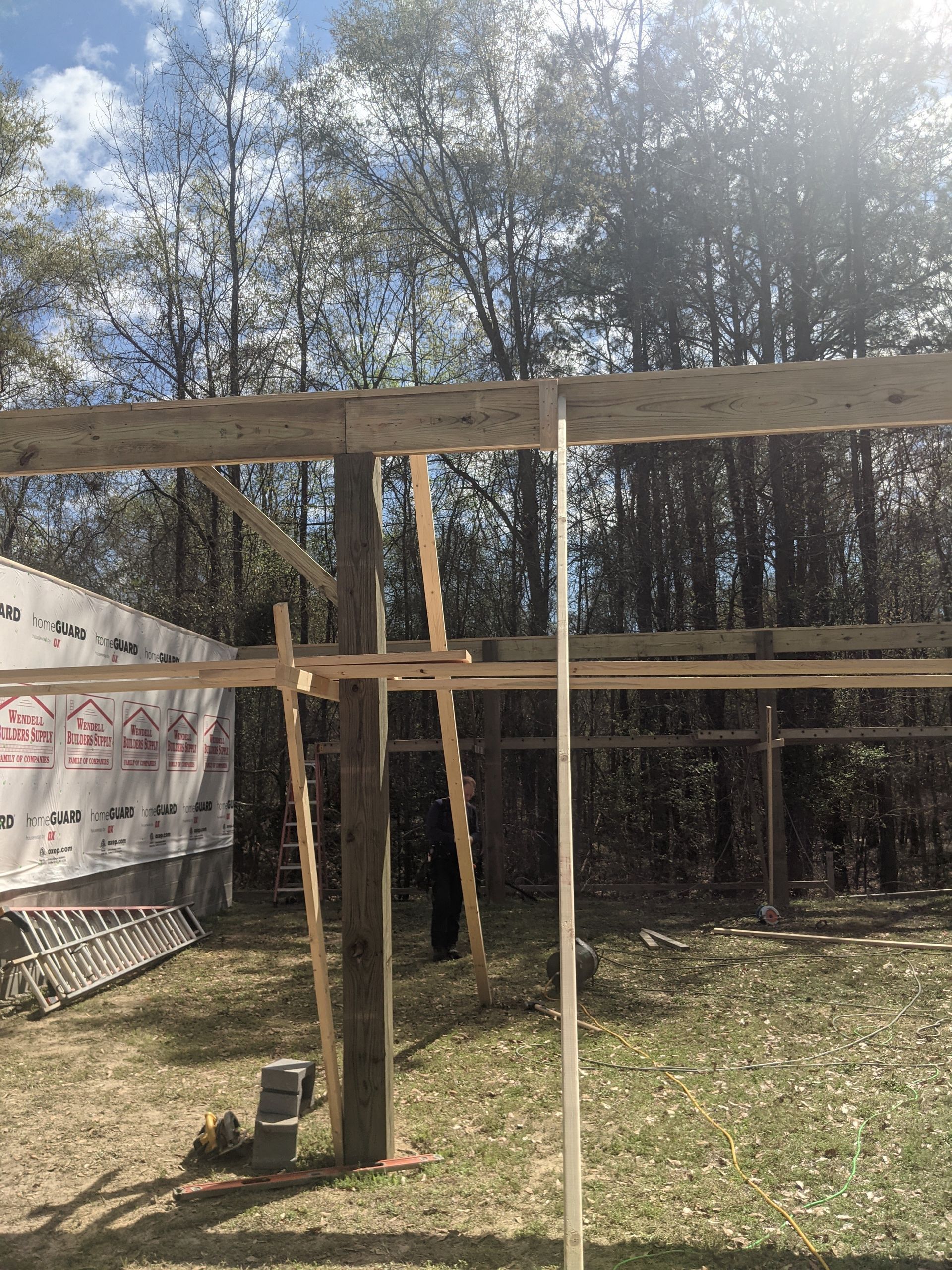 A wooden frame construction in progress, with beams and vertical posts set against a backdrop of trees.